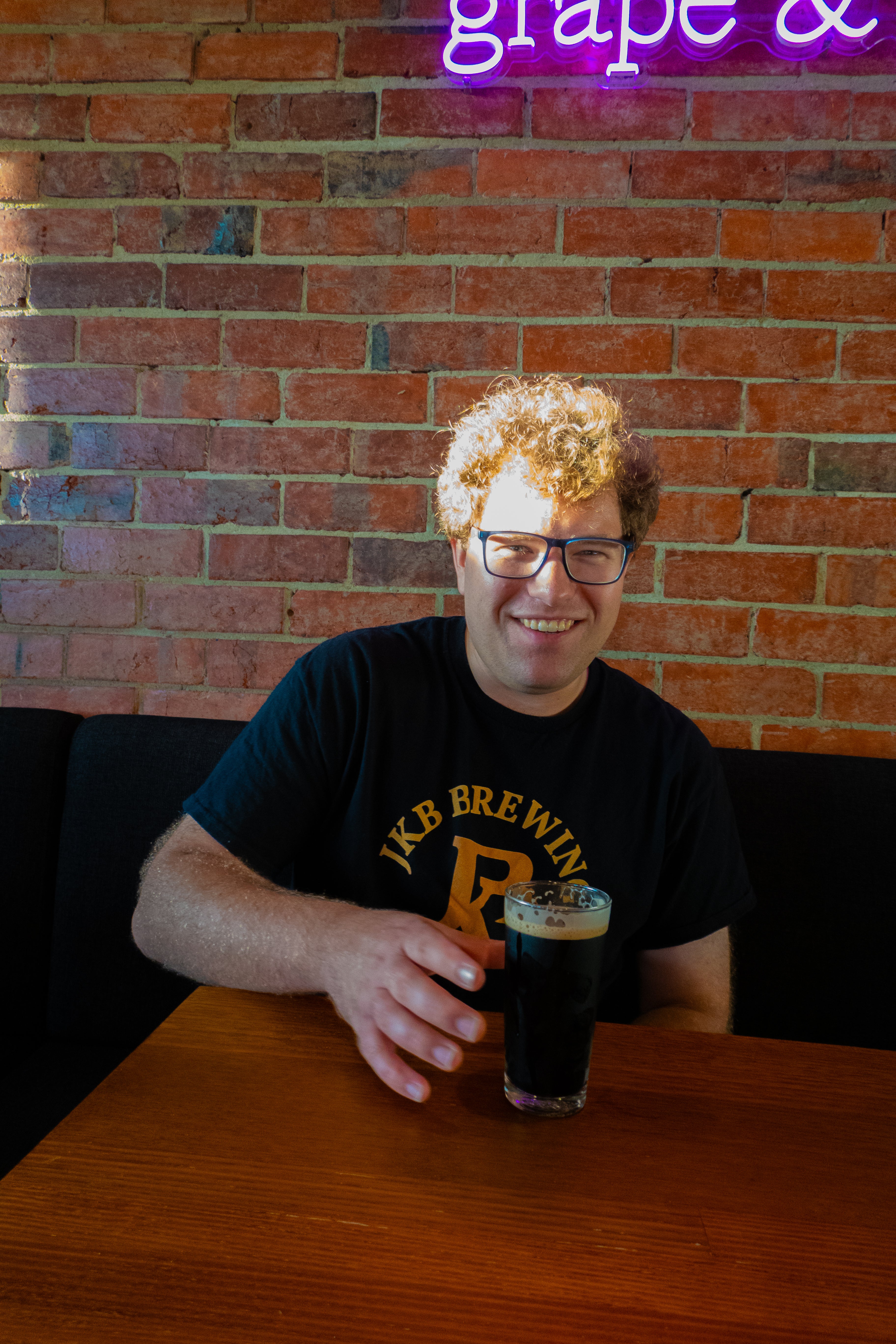 Person holding a glass of dark beer in front of a brick wall with a neon sign.
